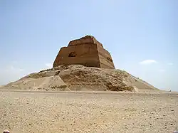 Fotografie pyramidy připomínající skálu stojící v poušti. Pyramida má viditelné tři stupně, první stupeň je největší, druhý je velký asi jako čtvrtina prvního a třetí stupeň je velký asi jako polovina druhého. Stojí na troskách svého bývalého obložení.