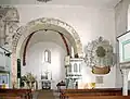 Pohled do interiéru, bílé stěny, levé dvě třetiny zaujímá vítězný oblouk s pohledem do kněžiště, na jeho pravém okraji světlá křtitelnice, vedle ní na zdi bohatě zdobený epitaf.