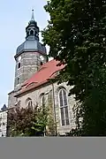 Stěna kostela z tmavého kamene s vysokými, obloukem zakončenými okny, vlevo vysoká věž s kopulí a špicí