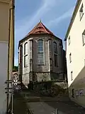 Šedohnědý chór s opěráky a vysokými okny, střecha červená s vikýři, krytá pálenými taškami, po stranách jsou budovy, mezi nimi dlážděná cesta se schody vedoucí ke kostelu