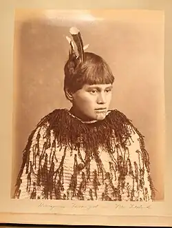 Maorská dívka, Horonyairu, poses against a plain background. She has short hair decorated with feathers and facial tattoos around