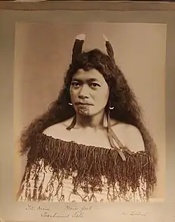 Maorská dívka, Titia Wikum (?), poses against a plain background. She has shoulder length hair decorated with feathers