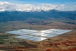 Parabolické žlaby pro koncentrovanou sluneční energii zářící na rovné pláni se zasněženými horami v pozadí.