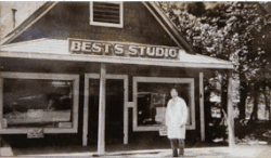 Na černobílé fotografii je muž oblečený jako malíř, stojící před dřevěným domem, na kterém je cedule s nápisem BEST´S STUDIO.