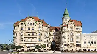Velká budova s výraznou rohovou věží zakončenou zelenou špičatou střechou. Budova má béžovou fasádu a červenou střechu, několik podlaží s řadou oken a balkónů. Architektura kombinuje secesní prvky s tradičnějšími styly. V přízemí jsou obchody nebo restaurace s venkovním posezením. Před budovou je otevřené prostranství. Fotografie je pořízena za jasného dne s modrou oblohou.