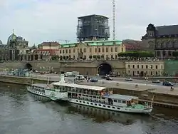Barevná fotografie s pohledem na Labe s výletní lodí a břeh s městskou zástavbou, ze které ční rozestavěný kostel Frauenkirche s nástavcem z lešení
