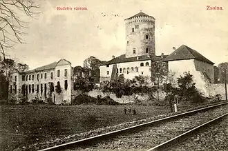 Sépiově nažloutlá černobílá pohlednice s hradem s velkou věží uprostřed. V popředí fotografie vlevo je pole, vpravo násep s jednokolejnou železniční tratí. Levá část fotografie je nadepsaná Budetin várrom, pravá Zsolna.