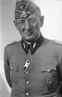 Max Simon jako SS-Standartenführer. V jeho druhé horní knoflíkové dírce můžeme vidět stuhu od železného kříže II.třídy se sponou. Na jeho náprsní kapse je železný kříž I. třídy a pod ním odznak za zranění v černém.