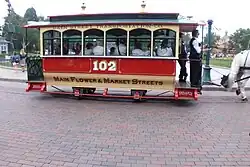 Tramvajový vůz červené barvy číslo 102 na dlážděné ploše. V pravé části zadek koně.