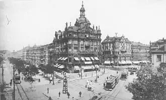 Černobílá fotografie zachycující nároží historizujícího domu s věžičkou obklopeného silnicemi, po kterých jezdí tramvaje