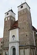 Barevná fotografie s pohledem na průčelí kostela se dvěma mohutnými věžemi na čtvercovém půdoryse