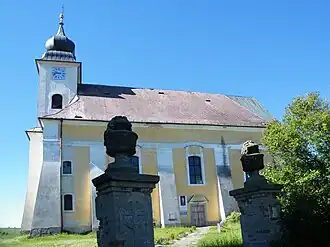 Osluněná jižní strana kostela s okny a dolů se zhmotňujícími opěráky, na levé straně kostela je kostelní věž, v popředí je otevřená vstupní brána