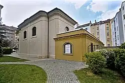 přední část kostela, hlavní vstup, stromoví a oplocení, v pozadí několik okolních domů