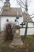 Barevná fotografie kovového kříže s kovovým podstavcem z roku 1866, který stojí u brány na hřbitov a ke kostelu