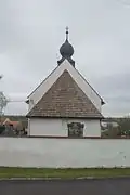 Barevná fotografie kostela při pohledu z východu, viditelná je osa kostela s apsidou, lodí a věží. V popředí je zeď hřbitova.