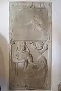 Barevná fotografie kamenného náhrobku ve zdi, v horní polovině je špatně čitelný nápis, ve spodní polovině je reliéf s ženskou postavou vlevo a se zabaleným dítětem a jeho duší vpravo, kterou má nad hlavou ve formě okřídlené hlavy. Mezi postavami je erb.