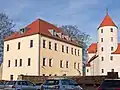 Barevná fotografie s pohledem na obdélníkovou budovu se sedlovou střechou v sousedství zámku