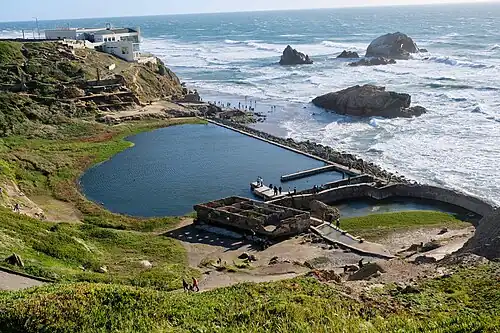 Pohled na zbytky lázní s domem Cliff House na útesu v pozadí, vpravo kamenný útvar Seal Rocks (2018)