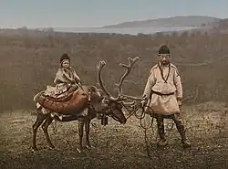 Dítě sedící na sobu, vpravo muž držící soba na uzdě. Oba v tradičním oblečení, s čepicemi.