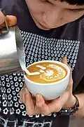 Barista vytvářející svou značku: anděla surfujícího na vlnách (šálek nedrží za ouško, protože je to příliš limitující při otáčení šálku, které je při technice latteart na vrcholové úrovni nezbytné).