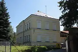 Boční pohled na objekt bývalé školy. Její patrová žlutě zbarvená budova stojí na mírném vršíčku. V popředí je veden pletivový plot.
