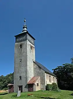 Vysoká věž kostela, za ní jednolodní kostel. Vše z šedivě zbarveného zdiva. Kolem kostela posekaná tráva, ze které vpravo na snímku vystupuje náhrobní kámen. V pozadí jsou vzrostlé listnaté stromy a nad celou scenérií je nebe zbarvené do sytě tmavě modré.