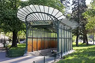 Zelený secesní vchodový pavilon do stanice pařížského metra s kovanou konstrukcí připomínající organické rostlinné tvary, se svítidly ve tvaru poupat a nápisem "METROPOLITAIN"
