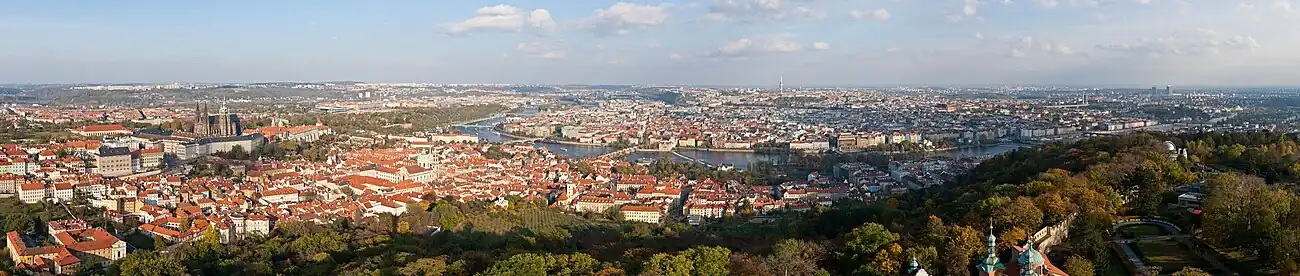 Panorama Prahy z Petřínské rozhledny. Vlevo jsou vidět Hradčany s Pražským hradem, jemuž dominuje svatovítská katedrála, pod nimi se rozkládá Malá Strana. Ve středu snímku pražský záhyb Vltavy obklopuje Staré Město. U horizontu leží Severní Město, Letná a Holešovice, Karlín, Vinohrady, Nové Město, Žižkov, Pankrác, Podolí i další čtvrti. V popředí snímku jsou zahrady vrchu Petřína, vpravo Růžový sad s věžemi starokatolického katedrálního kostela svatého Vavřince, Hladovou zdí, hvězdárnou a horní stanicí lanovky.
