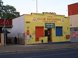 Zděný přízemní domek. Jeho čelní fasáda je žlutě zbarvená. V středu se nacházejí dveře a od nich nalevo okno s oranžovou roletou a napravo okno s modrou roletou. Nad vchodem je nápis „Bazar – zastavárna“. Levá boční fasáda objektu je bíle zbarvená