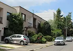 Po levé straně stojí řada patrových domů s rovnou střechou. Před nimi jsou malé zahrádky přerušované vjezdy do garáží. Na jednom z nich parkuje automobil peugeot. Nalevo je asfaltová vozovka, na níž parkují osobní automobily. Mezi předzahrádkami a vozovkou se nachází asfaltový chodník.