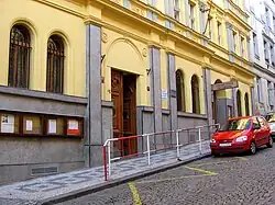 Vstup do zděného domu v kopci. Před ním je chodník a dlážděná silnice se zaparkovaným automobilem. Chodník do komunikace odděluje zábradlí.