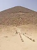 Pohled na pyramidu zespodu. Přibližně uprostřed stěny pyramidy se nachází otvor, k němuž vedou schody. Stojí u nich velbloud se třemi muži.