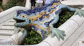 Mozaiková socha ještěra v Parku Güell v Barceloně. Socha je umístěna na bílém mozaikovém zábradlí nebo zdi a je vyzdobena barevnými keramickými úlomky v modré, žluté a zelené barvě. Tělo ještěra je modré s výraznými žlutými vzory, hlava a končetiny jsou zelené. Socha má otevřenou tlamu a je zachycena v pohybu po nakloněné ploše. V pozadí jsou vidět schody a část parku se zelení.