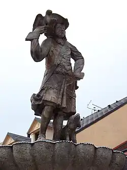 Pohled na sochu stojícího sokolníka v dobovém kroji s kloboukem. V pravé pokrčené ruce drží dravého ptáka a u levé nohy mu sedí lovecký pes. Částečně viditelná je i mušle, ve které je socha usazena.