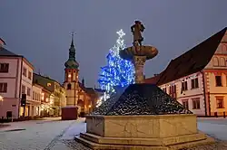 Vánoční pohled na zasněženou kašnu se sokolníkem s dřevěným bedněním. Napravo je budova bývalé radnice, za kašnou je rozsvícený Vánoční strom. V pozadí je vidět kostel svatého Jakuba Většího.