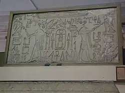 Kamenná stéla visící na zdi, na které se nacházejí dva polonazí muži s vlčí hlavou a sukní, jeden otočen na levou stranu a druhý na pravou. Oba mají pozvednuté levé ruce, muž na levé straně na levou stranu a muž na pravé straně na pravou stranu a drží v nich anchy. Druhá ruka obou mužů taktéž drží anch, ale není pozvednutá. Kolem mužů se nachází hieroglyfy a symboly
