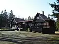 Podlouhlá přízemní budova tmavě hnědé barvy, která má po obou stranách obytné podkroví, před budovou terasa a štěrková plocha, za ní smrkový les