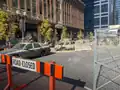 Ulice se špinavými, poškozenými auty a rozbitým betonem za oranžovočerným nápisem "Silnice uzavřena".