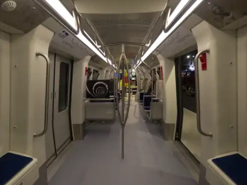 Interiér vozu metra, po obou stranách jsou vidět dveře, nad nimi orientační diagramy a plánky. Vlak má moderní design v bílé a šedé barvě, podlaha je světle modrá, rovná. Sedadla jsou uspořádána podél stěn, v barvách modré a bílé. Prostor je osvětlený zářivkami u stropu. Ulička mezi sedadly je prostorná, vhodná i pro stojící cestující. Je vidět řada kovových madel pro držení se za jízdy.
