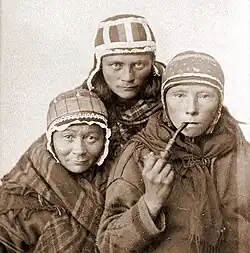 Černobílá fotografie tří sámských žen v pokrývkách hlavy, jedna kouří dýmku