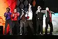 Six men dressed in variously colored clothes performing on a stage.