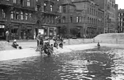 Varšava července 1944, měsíc před varšavským povstáním: Děti se koupou v hasičské vodní nádrži na Napoleonově náměstí. Za nimi Goldstanův městský dům na Napoleonově náměstí 5 / Moniuszki 1. Dále viditelná spodní patra budovy Prudentialu.