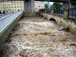 Barevná fotografie s vertikálním pohledem na rozvodněný přítok Labe, který dosahuje k horní části oblouku mostu