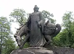 Socha muže ve zbroji a přilbou v ruce se svitkem, za ním stojí kůň se skloněnou hlavou. Sousoší je na skalnatém podstavci, v pozadí stromoví parku.